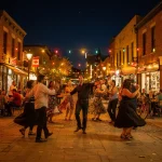 A vibrant evening street scene featuring a group of people dancing in the middle of a street adorned with colorful string lights. The street is lined with shops and outdoor cafes, where people are sitting at tables, enjoying the festive and cheerful atmosphere. Some dancers are wearing dresses, adding to the lively ambiance.