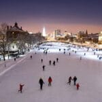 A vibrant winter scene featuring many people ice skating on a large outdoor rink surrounded by snow-covered trees and buildings. The city skyline is visible in the background, and the rink is illuminated by lights, suggesting it is evening.