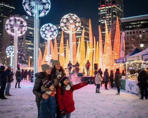 A lively outdoor scene at night featuring a family posing for a photo amidst decorative lights shaped like large dandelions and orange illuminated tree-like structures. A band performs on a stage as people gather around, and snow covers the ground. Illuminated buildings are visible in the background, contributing to the festive winter atmosphere.