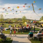 A vibrant outdoor scene in a park during a kite festival. The sky is filled with colorful kites of various shapes and sizes. A crowd of people, including children, adults, and individuals in wheelchairs, are enjoying the event. Some are gathered at picnic tables, others are seated on benches, and a playground is visible in the background. Tents and rolling green hills enhance the festive atmosphere under a partly cloudy sky.