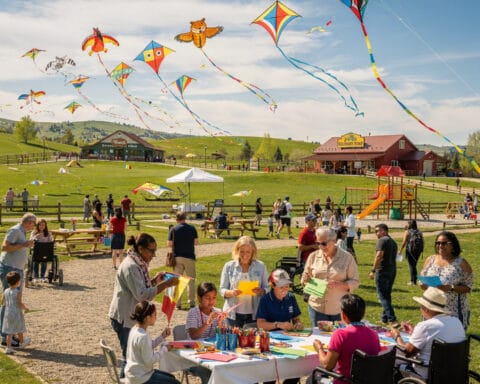 A lively outdoor scene of a kite festival with colorful kites of various shapes flying in a blue sky. People of all ages, including some in wheelchairs, are seated at tables doing crafts or drawing. The event is set in grassy fields with picnic tables and a play area featuring a slide. In the background, rolling hills and buildings enhance the festive community atmosphere.