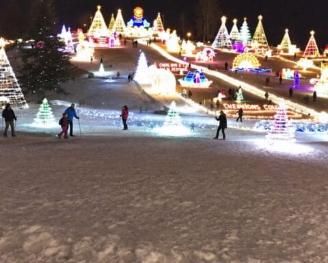 A snowy nighttime landscape featuring numerous illuminated holiday light displays in various shapes, such as trees and festive decorations. People are walking around, enjoying the vibrant and festive winter holiday atmosphere.