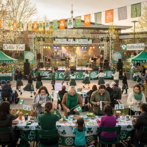 A lively outdoor St. Patrick's Day event with a Celtic theme at the Museum of Surrey. Musicians perform on a stage while people in traditional attire, including kilts, dance in front. Attendees sit at tables adorned with shamrock-themed decorations, engaging in crafts and enjoying the festive atmosphere. String lights and banners add to the colorful celebration.