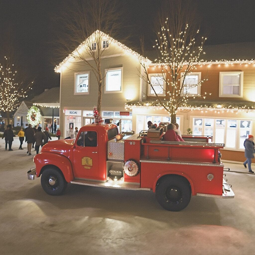 A nighttime festive scene featuring a vintage red fire truck decorated with holiday lights. The truck is parked in a pedestrian area surrounded by people enjoying the event. Nearby buildings and trees are adorned with holiday lights, and a large wreath is displayed on one of the buildings, creating a cheerful winter celebration atmosphere.