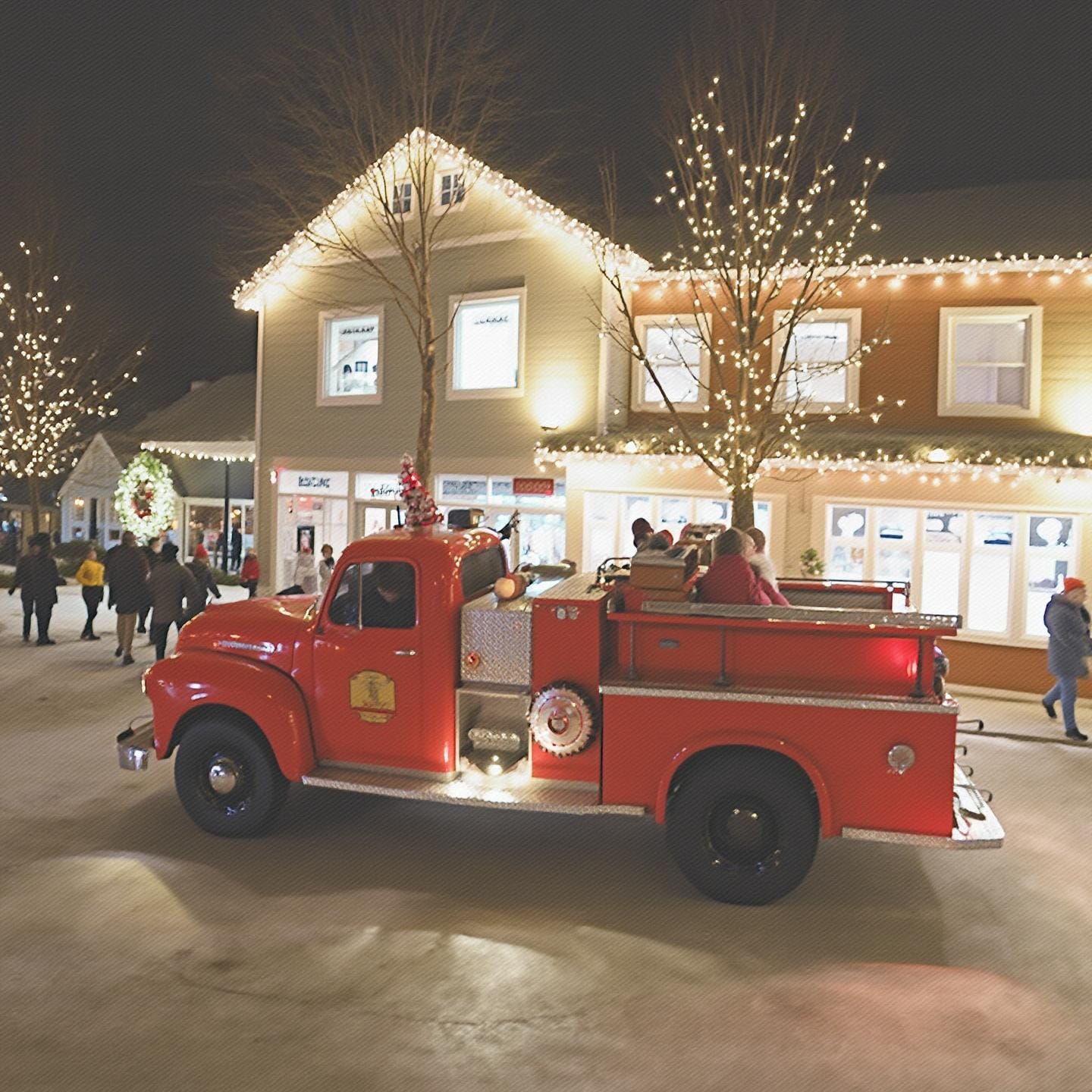 A nighttime festive scene featuring a vintage red fire truck decorated with holiday lights. The truck is parked in a pedestrian area surrounded by people enjoying the event. Nearby buildings and trees are adorned with holiday lights, and a large wreath is displayed on one of the buildings, creating a cheerful winter celebration atmosphere.