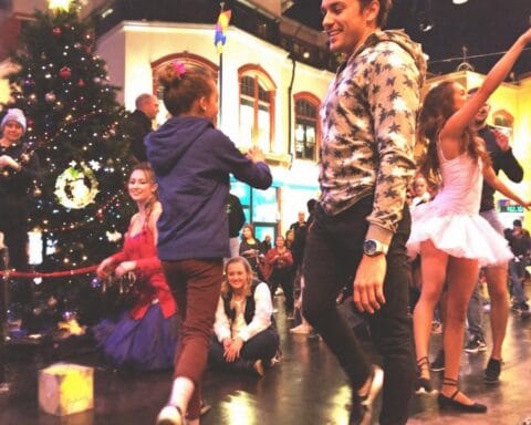 A lively outdoor scene featuring people dancing and celebrating near a decorated Christmas tree. The area is well-lit and set in an urban environment with a building in the background, creating a joyful and vibrant holiday atmosphere.