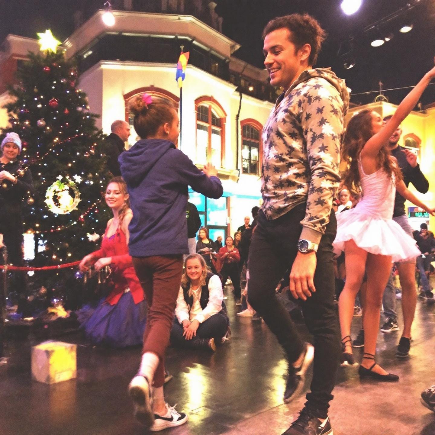 A lively outdoor scene featuring people dancing and celebrating near a decorated Christmas tree. The area is well-lit and set in an urban environment with a building in the background, creating a joyful and vibrant holiday atmosphere.