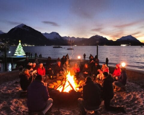 A group of people gathered around a bonfire on a sandy beach at dusk, with a mountainous background. A decorated Christmas tree with lights stands near the water in the distance, adding a festive and cozy ambiance to the serene scene.