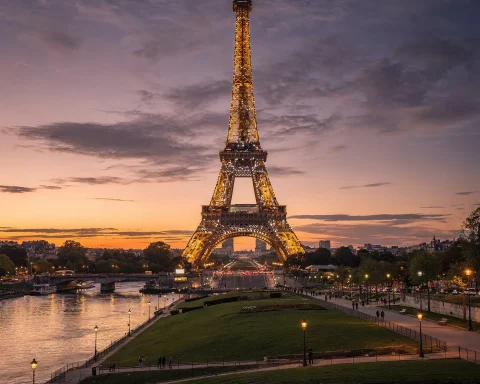 The Eiffel Tower in Paris, France, illuminated at dusk with a beautiful sunset in the background. A river flows in the foreground, and people are walking along green areas and pathways near the tower. The sky features a mix of clouds and a calming gradient of colors from the setting sun.