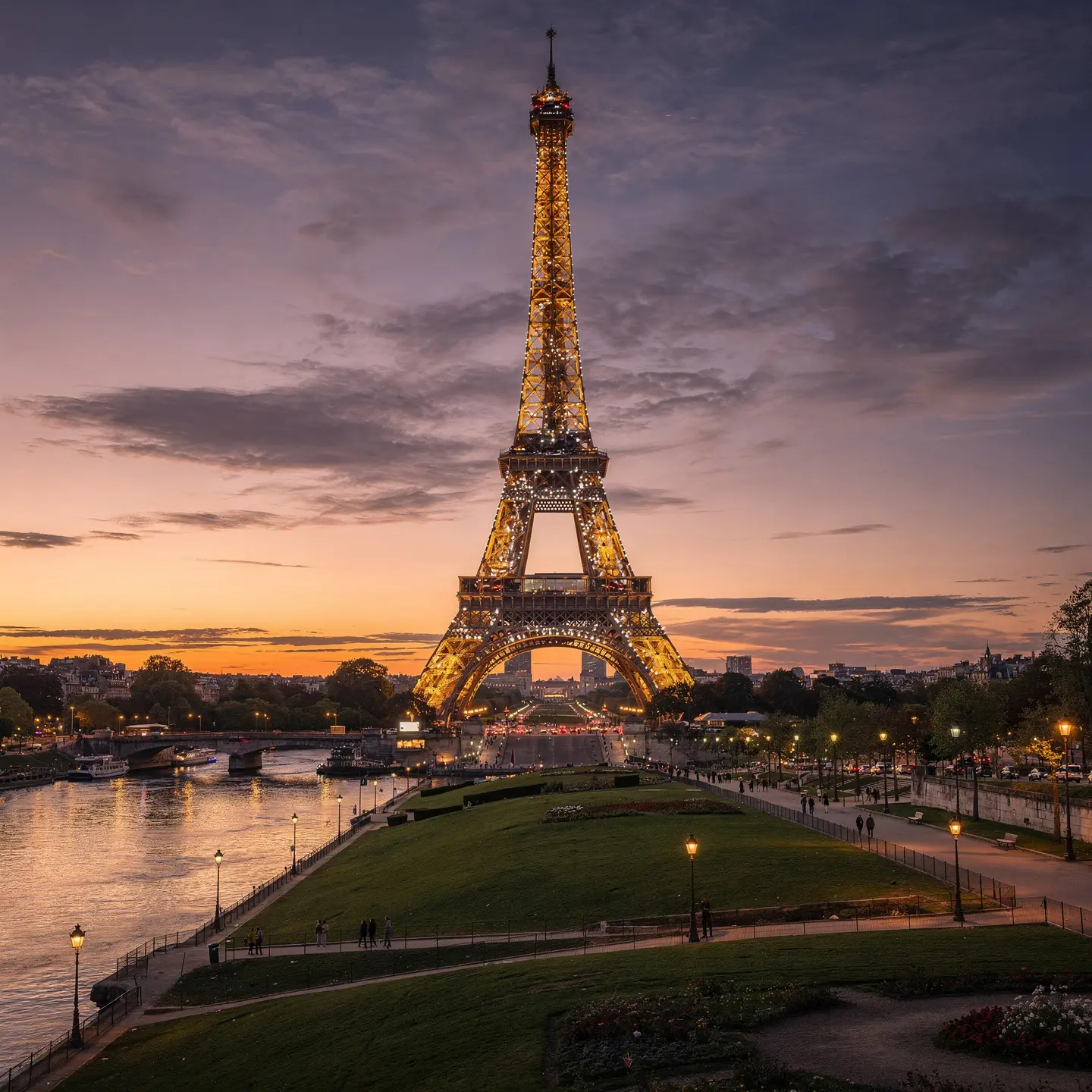 The Eiffel Tower in Paris, France, illuminated at dusk with a beautiful sunset in the background. A river flows in the foreground, and people are walking along green areas and pathways near the tower. The sky features a mix of clouds and a calming gradient of colors from the setting sun.
