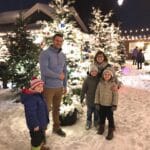 A family dressed in winter clothing stands in a festive, snowy setting surrounded by multiple decorated Christmas trees with bright lights and ornaments, creating a cheerful holiday atmosphere.