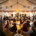 A lively scene inside a tent decorated with lights and flags, featuring people performing a traditional dance in detailed embroidered cultural costumes. A band is playing on a stage, enhancing the festive atmosphere of the cultural celebration.