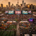 A lively outdoor FIFA Fan Festival event in Toronto, 2026, featuring large screens showing football matches, surrounded by a big crowd. The scene includes various tents, booths, a children's play area, a small sports area, and festive decorations with numerous flags. The Toronto city skyline with skyscrapers and the CN Tower is visible in the background.