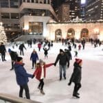 A festive ice skating rink set in a city environment, with people of all ages skating and enjoying the activity. A brightly decorated Christmas tree stands to the left, and lights adorn surrounding buildings, creating a cheerful holiday atmosphere. Tall high-rise buildings frame the background of the urban plaza.