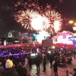 A large crowd gathers at night for an outdoor event with a vibrant fireworks display lighting up the sky. Colorful lights and structures enhance the festive atmosphere, with visible signs displaying the word 'metro' and people celebrating or enjoying the public event.