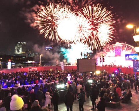 A large crowd gathers at night for an outdoor event with a vibrant fireworks display lighting up the sky. Colorful lights and structures enhance the festive atmosphere, with visible signs displaying the word 'metro' and people celebrating or enjoying the public event.