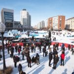 A snowy outdoor scene featuring a large group of people gathered in a public space surrounded by buildings. A stage is set up for a performer or band in the center, with attendees dressed in winter clothing indicating cold weather. Some people are standing, others are walking or sitting, and several ice structures are visible, creating a festive atmosphere suggestive of a winter festival or concert.