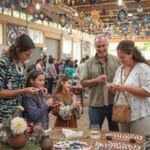 A festive event with a group of people engaged in crafting activities. Colorful decorations hang from the ceiling, and tables are set up with craft materials. The atmosphere is lively and community-focused, with participants enjoying themselves as they create and interact.