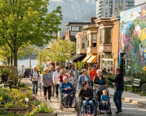 A lively street scene featuring a diverse group of people, including individuals walking and in wheelchairs. The street is lined with colorful buildings and storefronts, with a vibrant mural on one wall. In the background, snow-capped mountains and a body of water create a picturesque setting. A garden with various plants lines the sidewalk, and the weather is clear and pleasant.