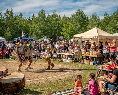 A lively cultural festival featuring performers in traditional Native American attire dancing in an outdoor setting. The dancers are surrounded by an audience, some seated and others standing, with tents and stalls visible in the background. A tent selling 'Fresh Frybread' and 'Bannock' is noticeable. The atmosphere is festive as attendees watch, take photos, and enjoy the event.