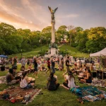 A vibrant outdoor scene featuring people sitting on the grass, playing drums, and engaging in various activities at a park. In the background, there is a tall monument topped with a statue. The atmosphere is lively, with stalls displaying items on blankets, and the setting sun casts a warm, golden light over the trees and gathering crowd.