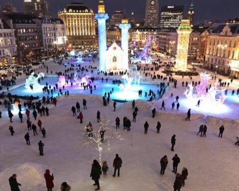 A snowy public square at night, lively with people walking and gathering near colorful illuminated decorations. The area features various lit sculptures and structures, surrounded by ornate buildings that enhance the festive atmosphere.