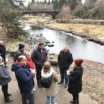 A group of people dressed in winter clothing stand and converse near a riverbank. In the background, a bridge spans across the river, and the area is surrounded by trees and grassy terrain, creating a natural and serene outdoor winter setting.