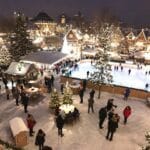 A lively Christmas market on a snowy winter evening, featuring an ice skating rink with people skating, brightly lit trees, and market stalls adorned with festive decorations. The area is bustling with people enjoying the cheerful holiday atmosphere.