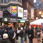 A lively street scene featuring a winter market with people gathered outside. Signs and banners are displayed, including one promoting 'Queer Craft Workshops' and another with a rainbow design reading 'LGBTQ Harbour.' The area is decorated festively with lights and a colorful-patterned white tent. People are dressed in warm clothing, suggesting a winter event or celebration.