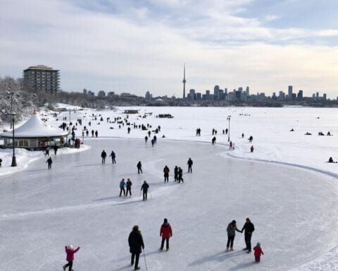 A winter scene showing people ice skating on a frozen body of water with a distant city skyline featuring a prominent tower in the background. The surrounding area is covered in snow, and many individuals are enjoying various outdoor activities on the ice. The location appears to be a popular recreational spot during the winter season.