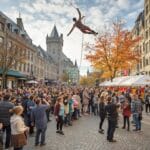 A vibrant street scene during a festival in an urban area with historic buildings. A performer is suspended in the air, performing an aerial act while a large crowd watches and takes photos. Trees with autumn foliage line the street, and vendor tents are set up along the side, adding to the festive atmosphere.