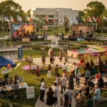 An outdoor festival showcasing multicultural activities and performances. The lively scene includes people enjoying a 'Jazz Stage' concert and another performance on the 'Rhythms of Canada' stage. Nearby, individuals in traditional Indigenous attire perform a dance. Food stalls offer dishes like butter chicken and poutine. Participants are engaging in activities such as Bollywood dance lessons and kite decorating. Various national flags are displayed, reflecting the event's cultural diversity. The setting features a garden area with bicycles parked nearby, enhancing the festive and welcoming atmosphere.
