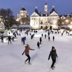 A lively outdoor ice skating rink with people of all ages enjoying the activity. In the background, a large, elegant building is lit up, along with a visible domed structure, creating a cheerful and festive winter atmosphere.
