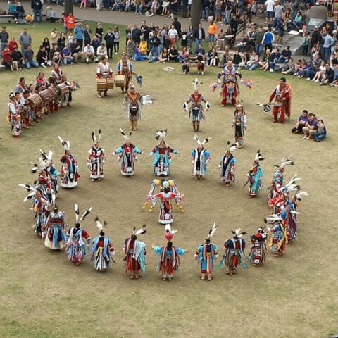 A group of people in traditional Native American attire performing a dance in a circle, accompanied by musicians playing drums. Spectators are gathered around the performance area, observing the cultural event.