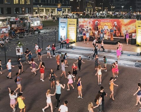A vibrant outdoor event featuring people dancing on a plaza with a stage and performers in the background. The scene includes string lights, food trucks, and groups of attendees dancing together, creating a festive and celebratory atmosphere.