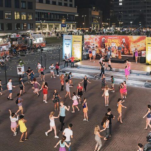 A vibrant outdoor event featuring people dancing on a plaza with a stage and performers in the background. The scene includes string lights, food trucks, and groups of attendees dancing together, creating a festive and celebratory atmosphere.