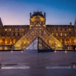 The Louvre Pyramid in Paris, France, illuminated during the evening. The large glass and metal pyramid serves as the main entrance to the Louvre Museum, surrounded by the historic Louvre Palace.