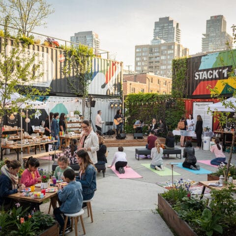 A vibrant outdoor event in an urban area featuring people painting at tables, practicing yoga on mats, and enjoying a live music performance. Booths displaying items are set up, and colorful shipping containers and greenery serve as the backdrop. The scene conveys a relaxed, communal vibe with participants engaging in various activities.