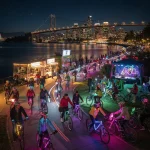 A lively night scene showcasing a group of cyclists riding along a waterfront path. The bicycles are decorated with colorful lights, creating a festive atmosphere. In the background, a brightly lit city skyline and a bridge can be seen spanning the water. Food trucks and a music performance setup with performers are visible by the path, adding to the energetic, celebratory mood of the scene.