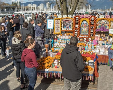 An outdoor altar for the Day of the Dead featuring marigold flowers, decorative skulls, and framed pictures. People are gathered around the display, observing and interacting, in a public park setting.