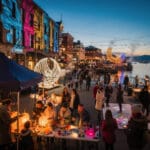A lively street festival by a waterfront at dusk, with people gathered at tables for arts and crafts activities. Buildings feature colorful light projections, decorative string lights are overhead, and sculptural light installations create a festive atmosphere. The scene captures a vibrant and social evening event.