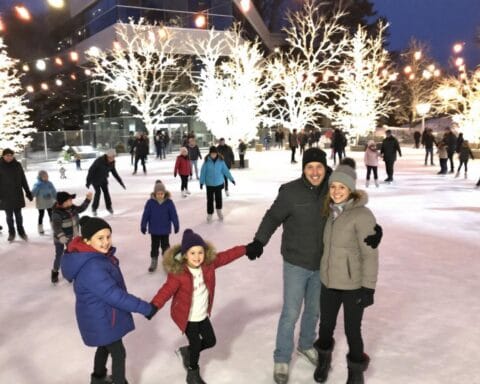 A group of people skating on an outdoor ice rink under strings of lights. Illuminated trees are visible in the background, creating a festive winter atmosphere. Everyone is enjoying the activity.