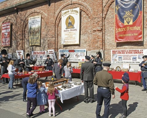 An outdoor event with people gathered around tables covered in red cloths. Uniformed individuals and a group playing bagpipes are present. Children and adults examine trophies and framed items on display. A brick wall in the background features large, colorful banners.