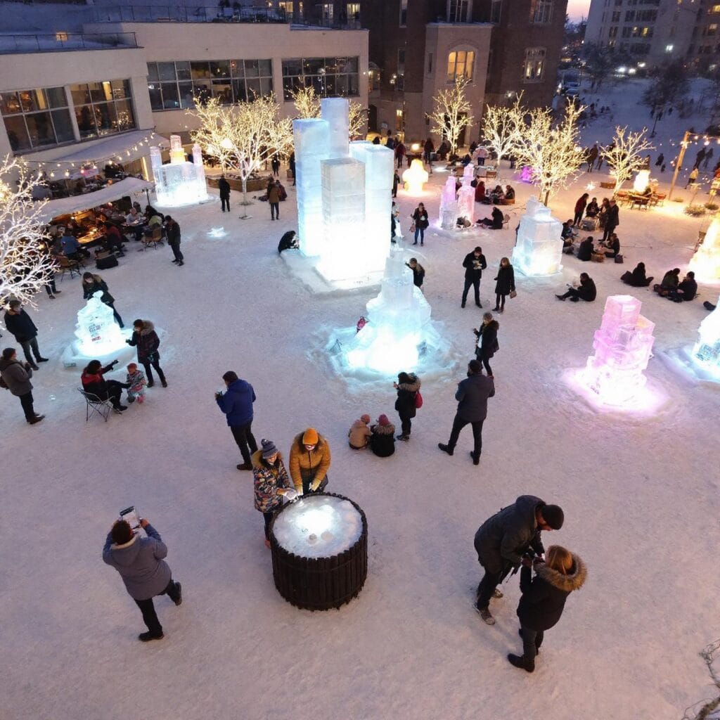 A winter scene featuring people gathered around illuminated ice sculptures of various shapes and sizes in an outdoor area covered with snow. Some individuals are sitting or standing near the sculptures, while others are taking photos or interacting with the environment. The area is decorated with lit-up trees and includes seating, creating a festive winter-themed atmosphere.