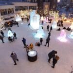 A winter scene featuring people gathered around illuminated ice sculptures of various shapes and sizes in an outdoor area covered with snow. Some individuals are sitting or standing near the sculptures, while others are taking photos or interacting with the environment. The area is decorated with lit-up trees and includes seating, creating a festive winter-themed atmosphere.