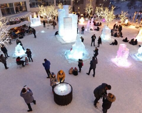 A winter scene featuring people gathered around illuminated ice sculptures of various shapes and sizes in an outdoor area covered with snow. Some individuals are sitting or standing near the sculptures, while others are taking photos or interacting with the environment. The area is decorated with lit-up trees and includes seating, creating a festive winter-themed atmosphere.